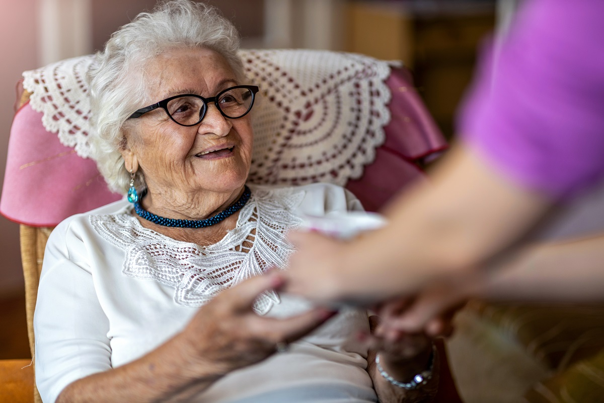 An elderly woman with white hair, glasses and a lace collar smiles as a care worker in a pink top offers her something.