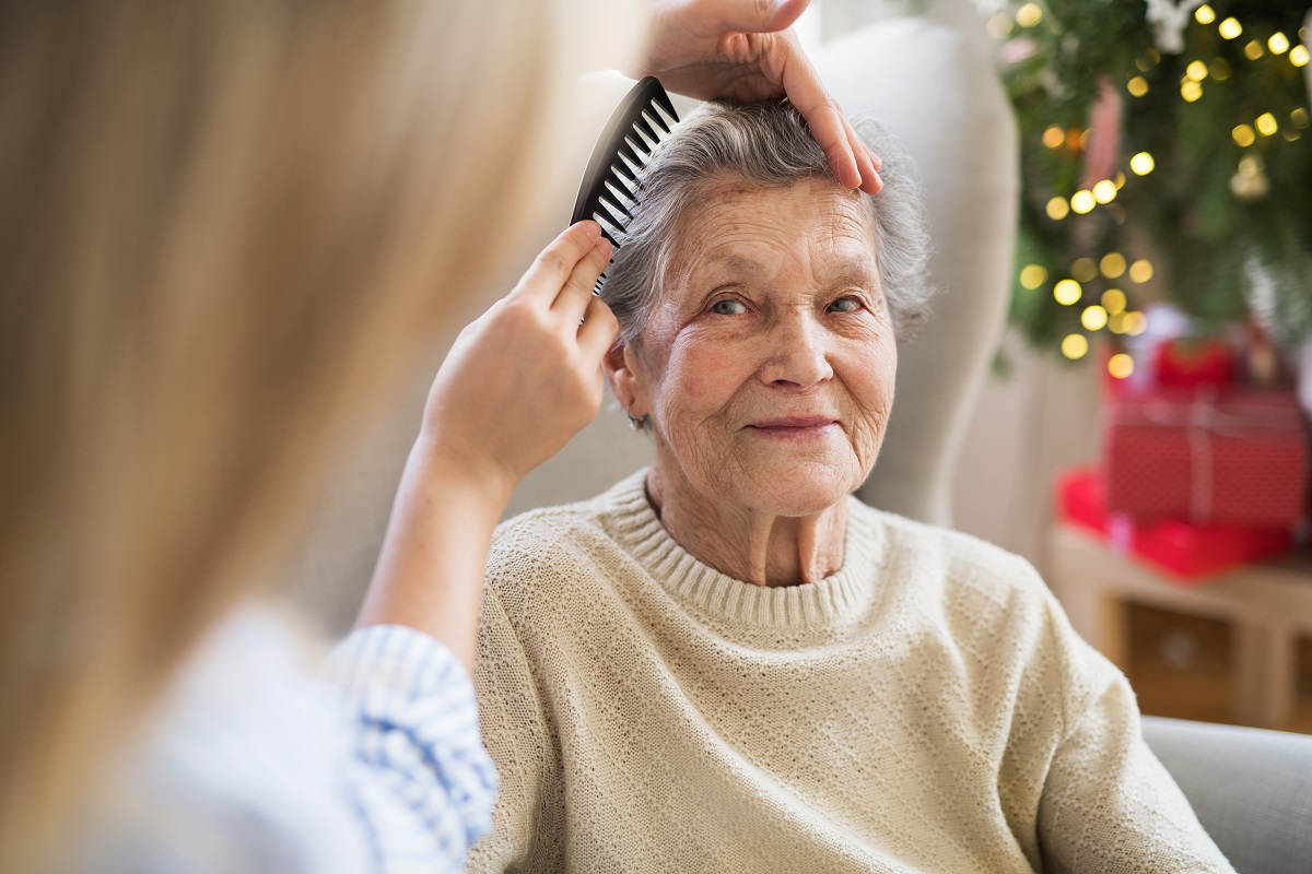 A younger person brushes the hair of an elderly woman with grey hair, who sits smiling near a decorated Christmas tree with gifts.