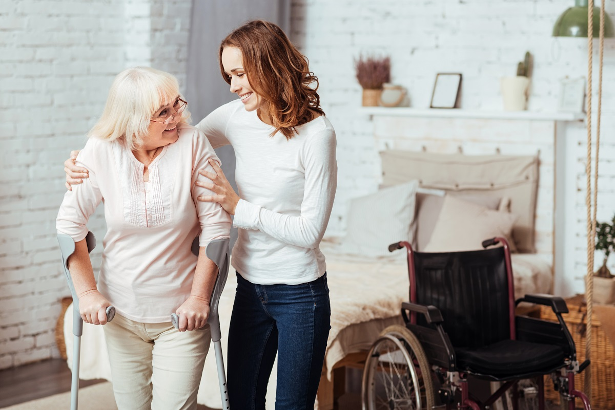 An older woman with white hair uses crutches to walk while a younger woman supports her arm, with a wheelchair nearby in a bedroom.