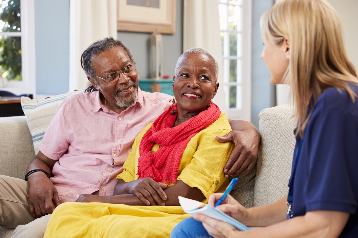 A couple sit together on a sofa talking with a care worker, who takes notes.