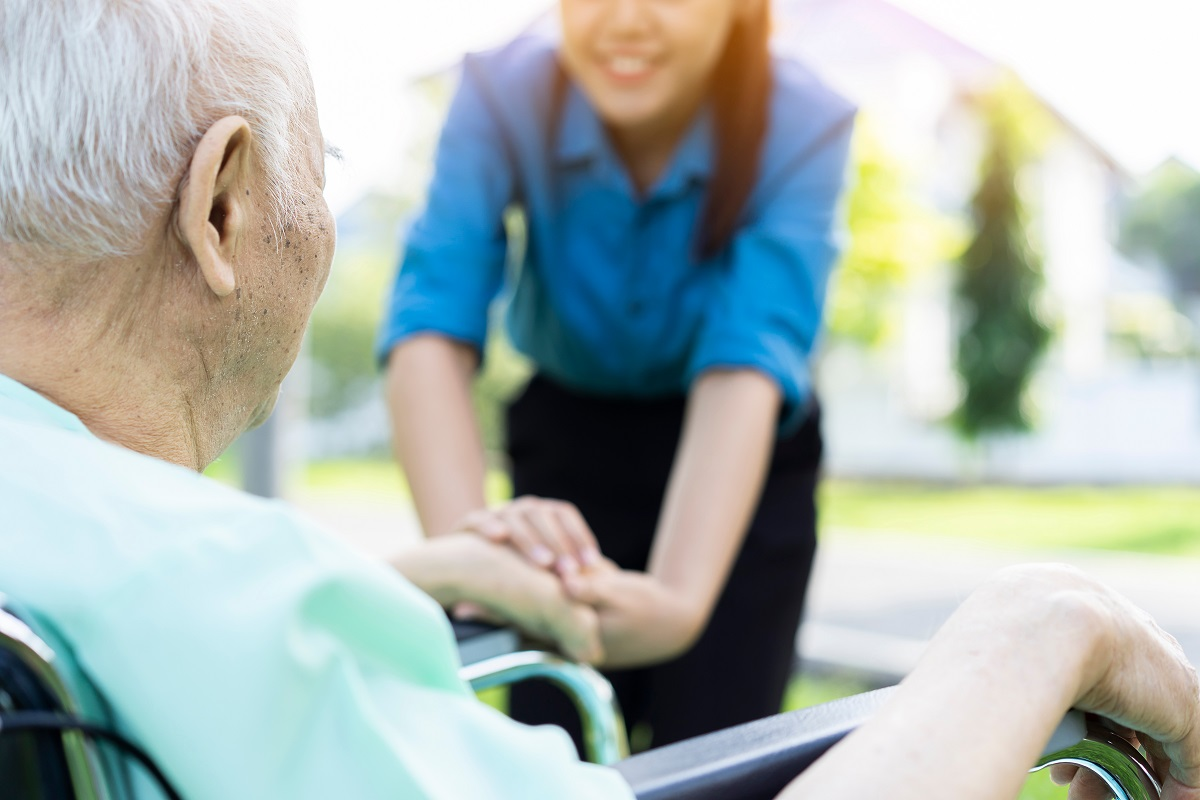 An older man in a wheelchair holds hands with a care worker outdoors.