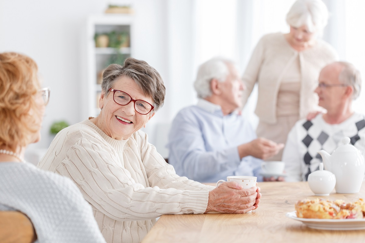 An older woman smiles while sitting at a table with other older adults enjoying tea and cake.