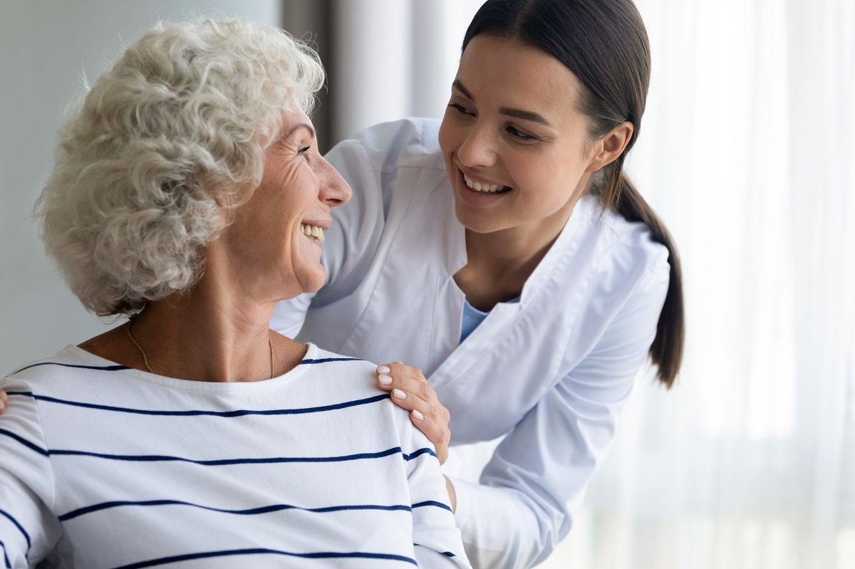 A young care worker smiling warmly at an older woman, gently resting a hand on her shoulder.