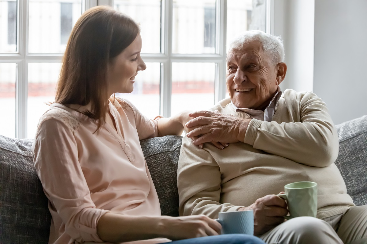 A younger woman rests a hand on the shoulder of an older man as they sit together on a sofa holding cups of tea.