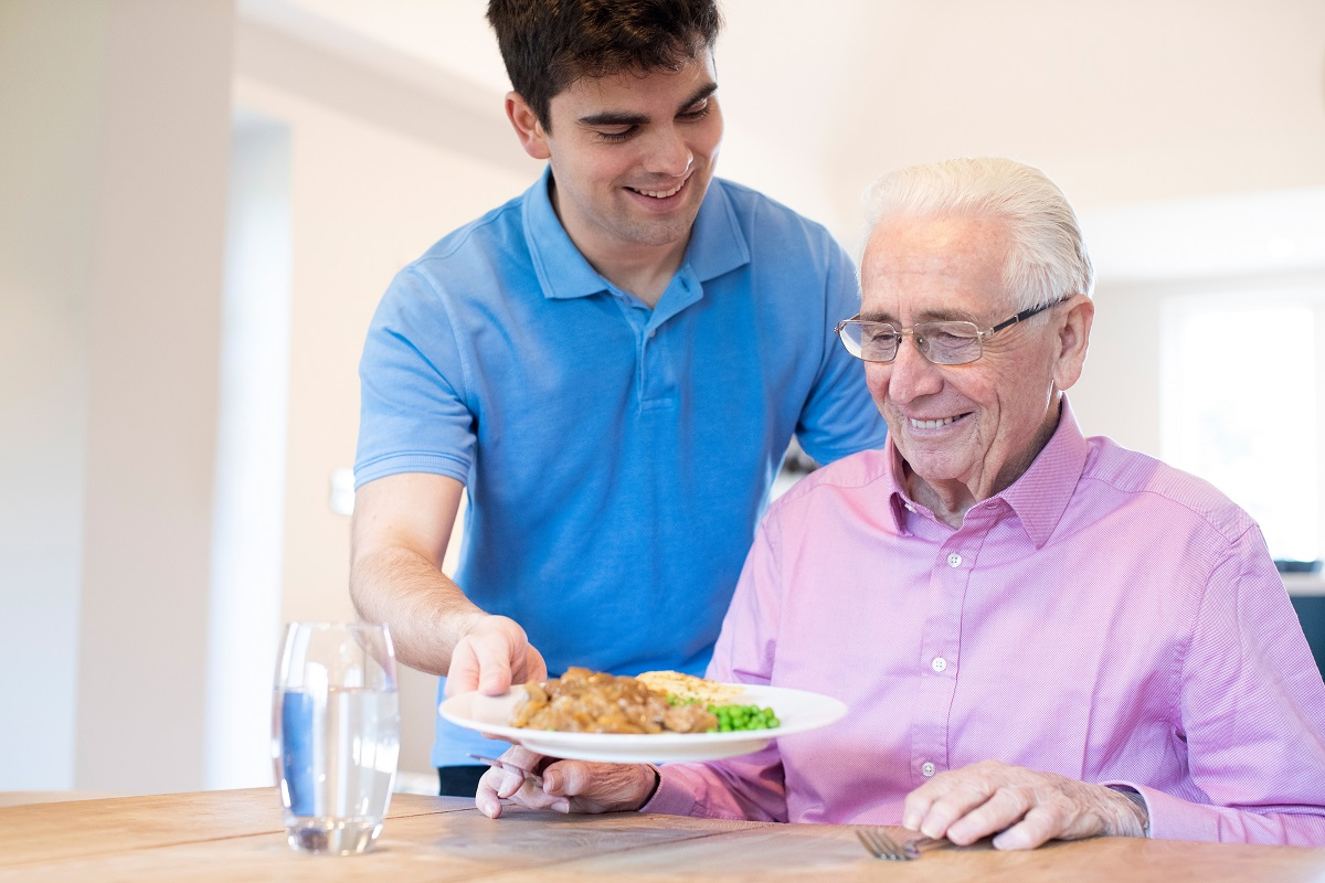 A young man in a blue polo shirt smiles as he serves a plate of food to an older man with white hair and glasses at a table.