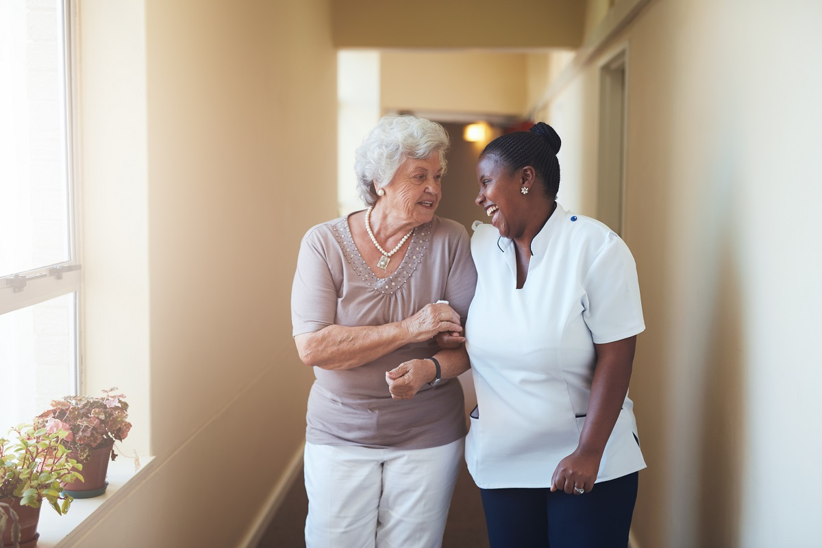 An older woman links arms with a smiling care worker as they walk together down a corridor.