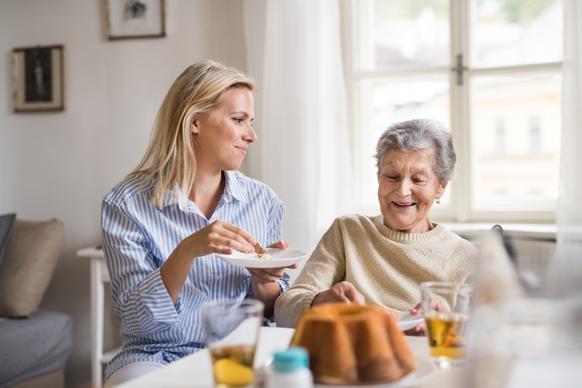 A younger woman in a striped top smiles while helping an older woman with grey hair eat at a table near a window.
