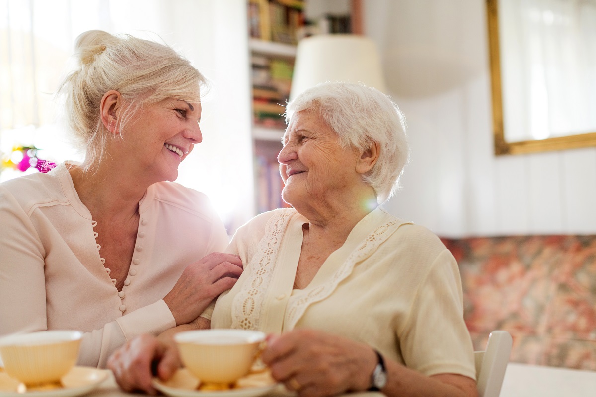 An older woman and a younger woman sit together smiling, holding cups of tea.
