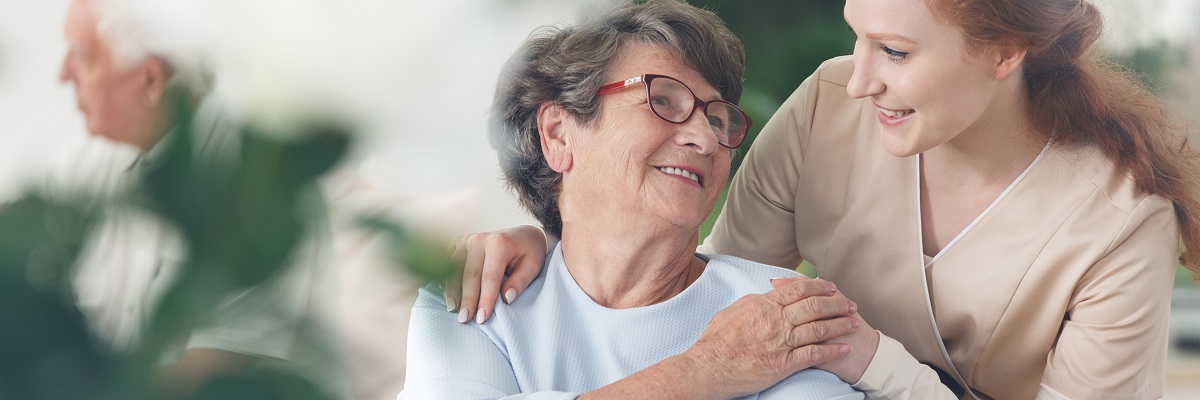 An older woman wearing glasses smiles as a younger woman rests a hand on her shoulder.