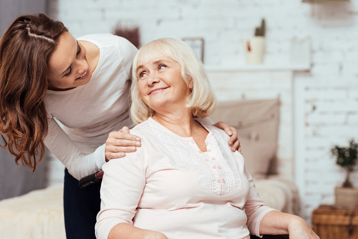 A smiling care worker rests a hand on the shoulder of a seated, smiling older woman in a bright bedroom.