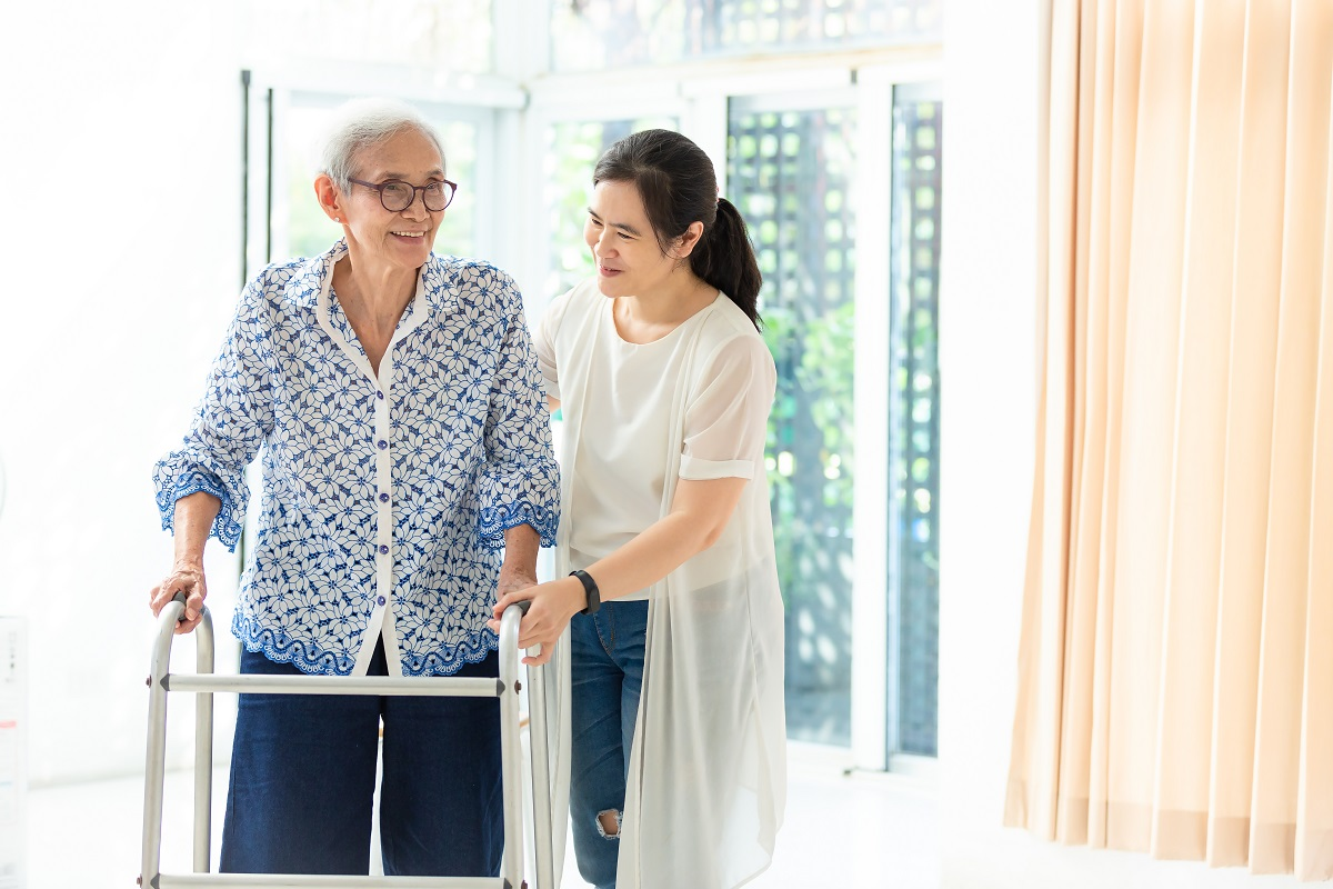 An older woman using a walking frame is helped by a younger woman.