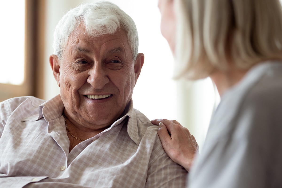 An older man smiles as a woman rests a hand on his shoulder.